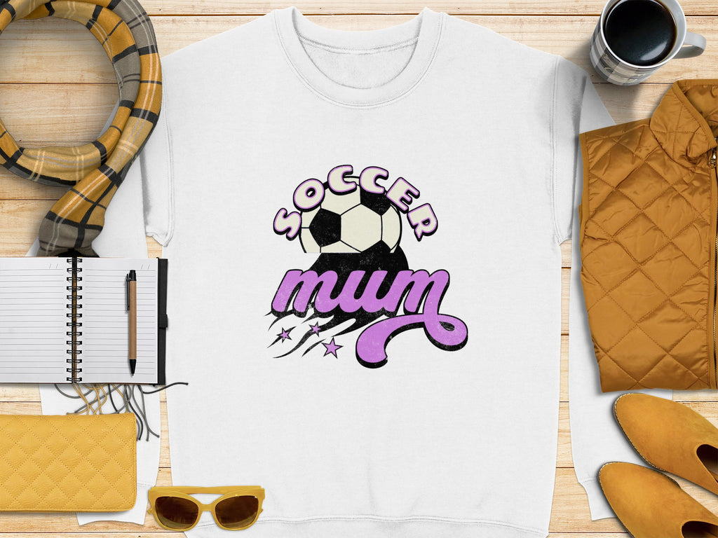 A Garment Graphics white sweatshirt with a vintage Soccer Mum design and soccer ball rests on a wooden surface. Nearby are a notebook with pen, yellow quilted purse, sunglasses, scarf, yellow jacket, and coffee cup.