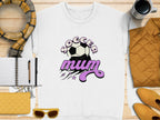 A Garment Graphics white sweatshirt with a vintage Soccer Mum design and soccer ball rests on a wooden surface. Nearby are a notebook with pen, yellow quilted purse, sunglasses, scarf, yellow jacket, and coffee cup.
