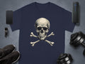 A dark graphic t-shirt by Garment Graphics, featuring a skull and crossbones design, is placed on a gray surface. Surrounding it are sleek accessories: smartphone, smartwatch, wireless speaker, black sneakers, and dumbbells.