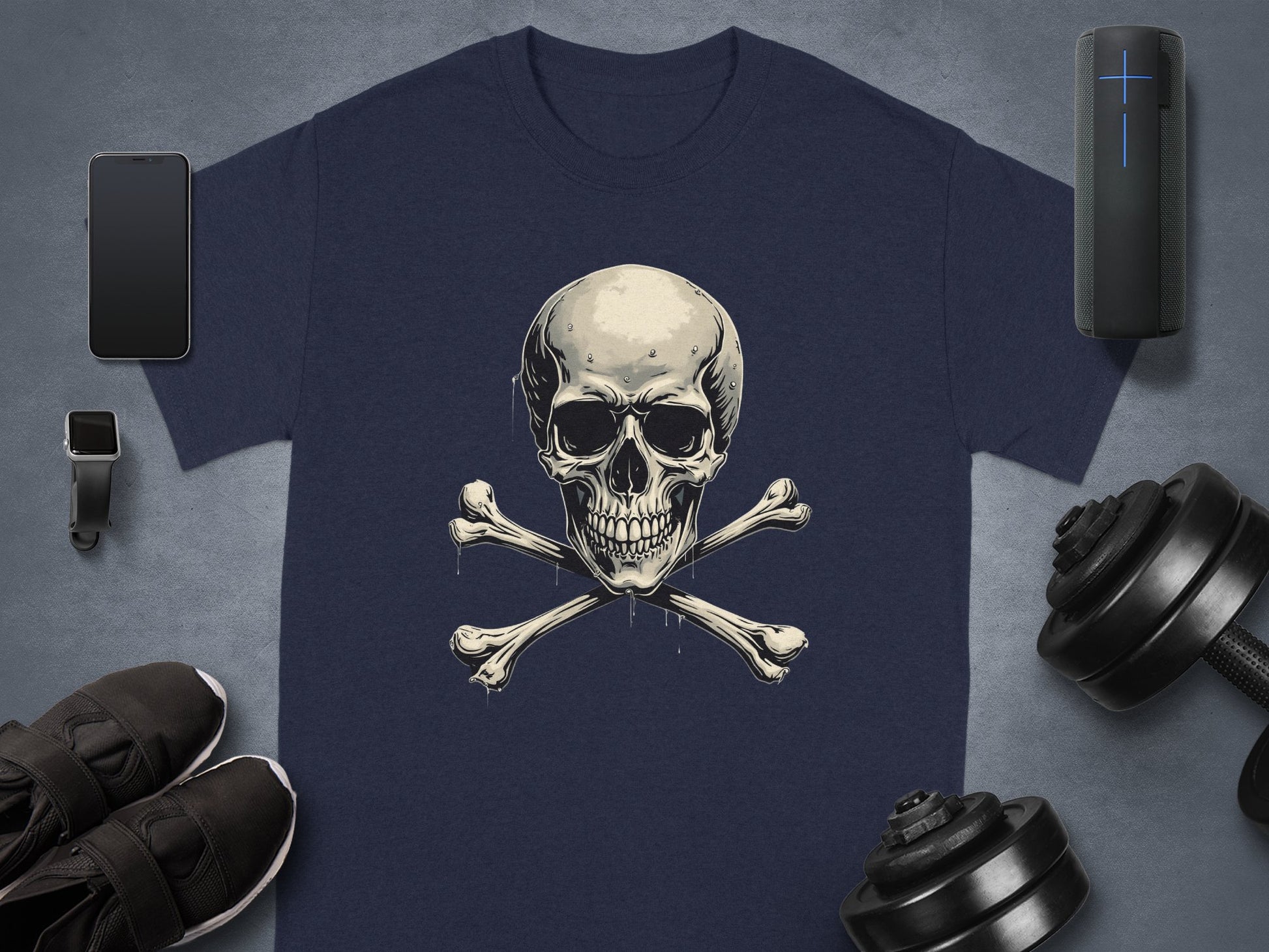 A dark graphic t-shirt by Garment Graphics, featuring a skull and crossbones design, is placed on a gray surface. Surrounding it are sleek accessories: smartphone, smartwatch, wireless speaker, black sneakers, and dumbbells.