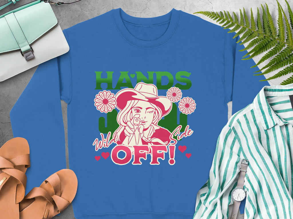 The Garment Graphics Wild Side Sweatshirt features a cartoon character in a hat with a flower, playful text, and hearts. Its loose fit complements casual items like striped shirts, sandals, bags, watches, and green leaves.