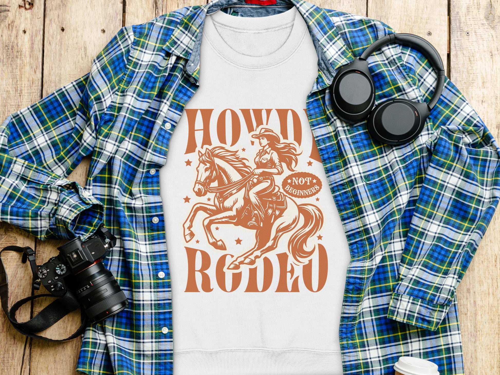 A flat lay image features a white unisex Garment Graphics T-shirt with a cowgirl on a horse and Howdy Rodeo text. Its layered on a plaid shirt. Next to it are headphones, a camera, and a disposable coffee cup on the wooden surface—ideal for graphic print sweatshirt enthusiasts!.