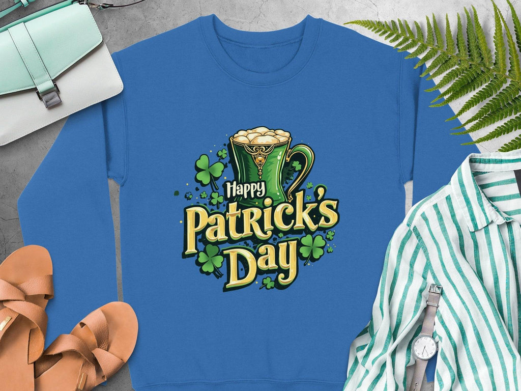 A vibrant sweatshirt from Garment Graphics featuring a Happy Patricks Day graphic with a green mug of frothy beverage and shamrocks, laid on a gray surface with sandals, a striped shirt, watch, purse, and nearby ferns.