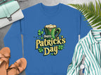 A vibrant sweatshirt from Garment Graphics featuring a Happy Patricks Day graphic with a green mug of frothy beverage and shamrocks, laid on a gray surface with sandals, a striped shirt, watch, purse, and nearby ferns.