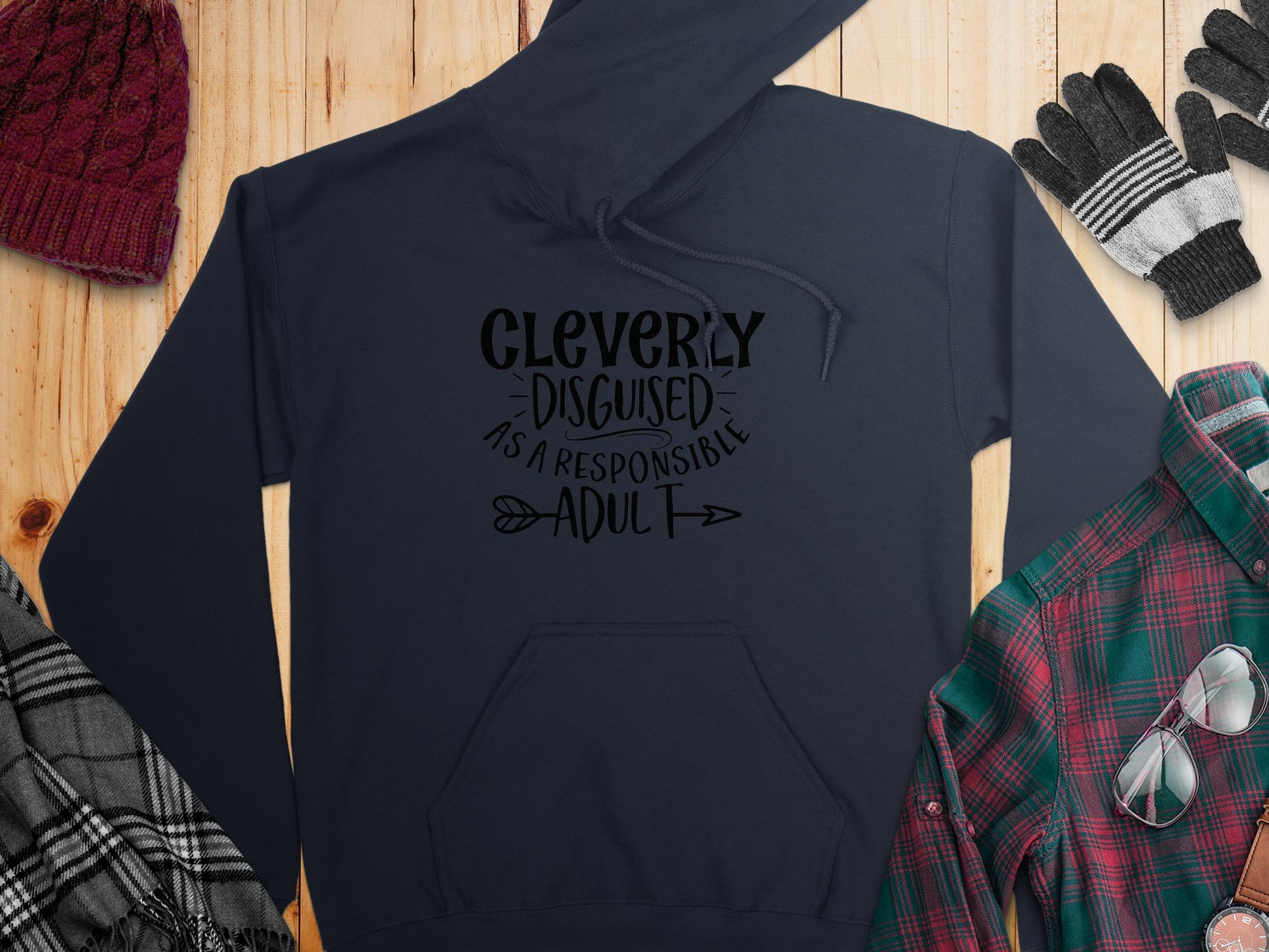 From Garment Graphics, the black hoodie in the Responsible Adult Hoodies collection reads Cleverly disguised as a responsible adult. It rests on a wooden surface with a red knit hat, fingerless gloves, gray plaid scarf, and green and red plaid shirt nearby.