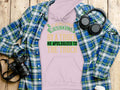 The Garment Graphics hoodie with the quote If an engineer gets run over by a truck, he apologizes to the truck is displayed on wood, surrounded by a blue plaid shirt, headphones, a camera, and a lidded cup—ideal for showcasing Englishmans manners stylishly.