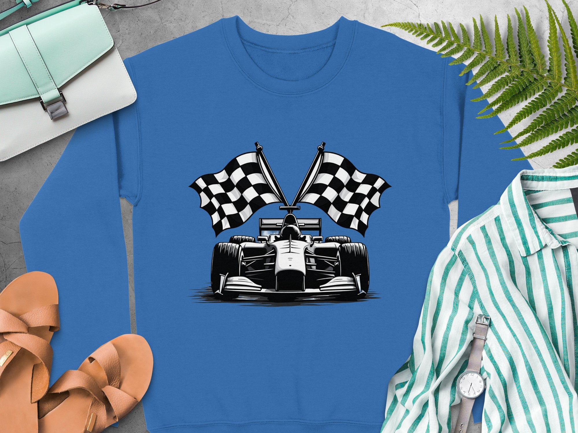 Garment Graphics blue sweatshirt features a racing car design with crossed checkered flags, styled on concrete with brown sandals, a white and teal purse, a plant, and complemented by a striped shirt with a watch.