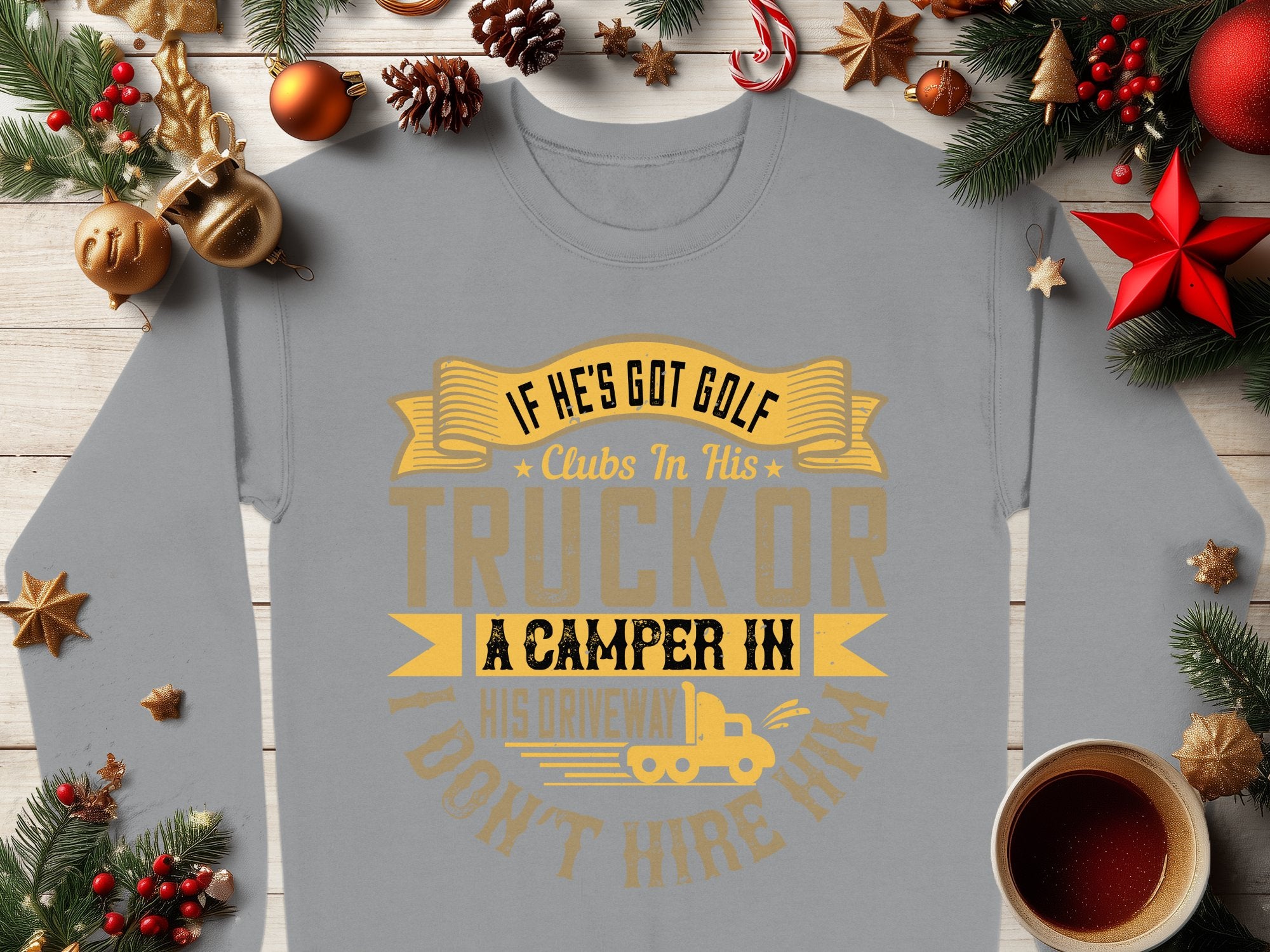 Garment Graphics offers a gray sweatshirt with the phrase If he’s got golf clubs in his truck or a camper in his driveway, dont hire him! in gold and yellow letters. Ideal for outdoor enthusiasts, its adorned with pine branches, ornaments, and coffee on a wooden table.