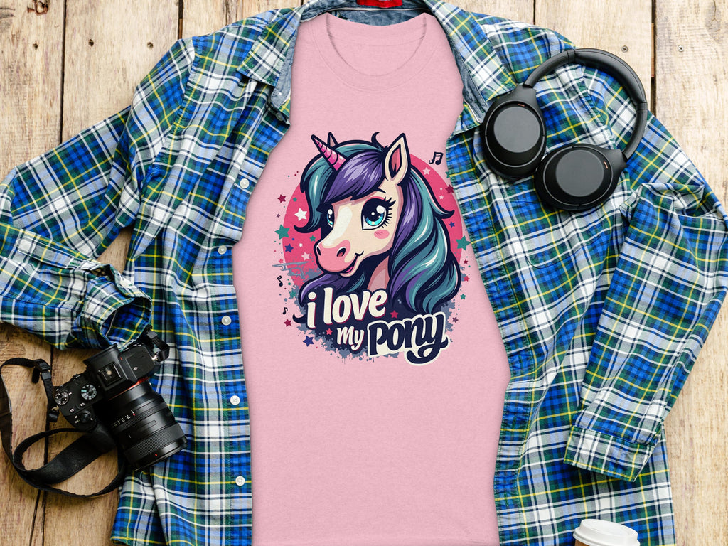 A Garment Graphics cartoon unicorn T-shirt with I love my pony is on a wooden surface, surrounded by a blue-green plaid shirt, headphones, a black camera, and a white cup—ideal for unicorn lovers.