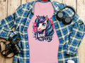 A Garment Graphics cartoon unicorn T-shirt with I love my pony is on a wooden surface, surrounded by a blue-green plaid shirt, headphones, a black camera, and a white cup—ideal for unicorn lovers.