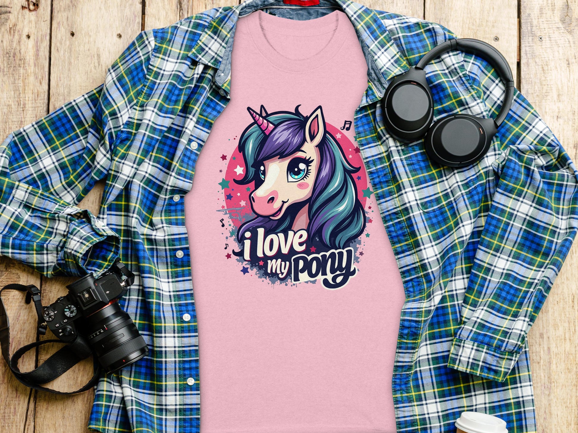 A Garment Graphics cartoon unicorn T-shirt with I love my pony is on a wooden surface, surrounded by a blue-green plaid shirt, headphones, a black camera, and a white cup—ideal for unicorn lovers.