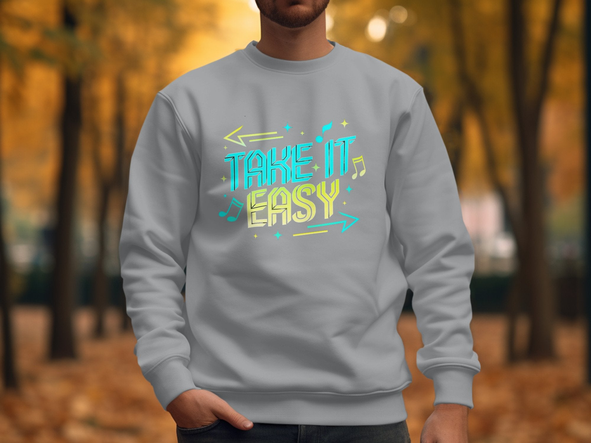 A person is wearing a Garment Graphics light gray sweatshirt with unisex sizing, featuring Take It Easy in vibrant letters. The backdrop shows a blurred tree-lined path with autumn leaves scattered on the ground.