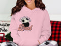 Lounging cozily, a person wears a pink Soccer Dad hoodie, featuring a soccer ball graphic, from Garment Graphics. Theyre seated with a festive pillow and wrapped in a red and black plaid blanket.