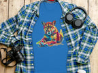 A Garment Graphics blue graphic T-shirt with a vibrant resting lion artwork is laid on a plaid shirt, alongside black headphones, a camera, and a white coffee cup, all arranged on the wooden surface.