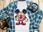 A quirky white T-shirt by Garment Graphics, featuring a cute cartoon mouse inside a donut, rests on a wooden surface. Nearby items for a playful wardrobe include a blue plaid shirt, headphones, a camera, and a white travel mug.