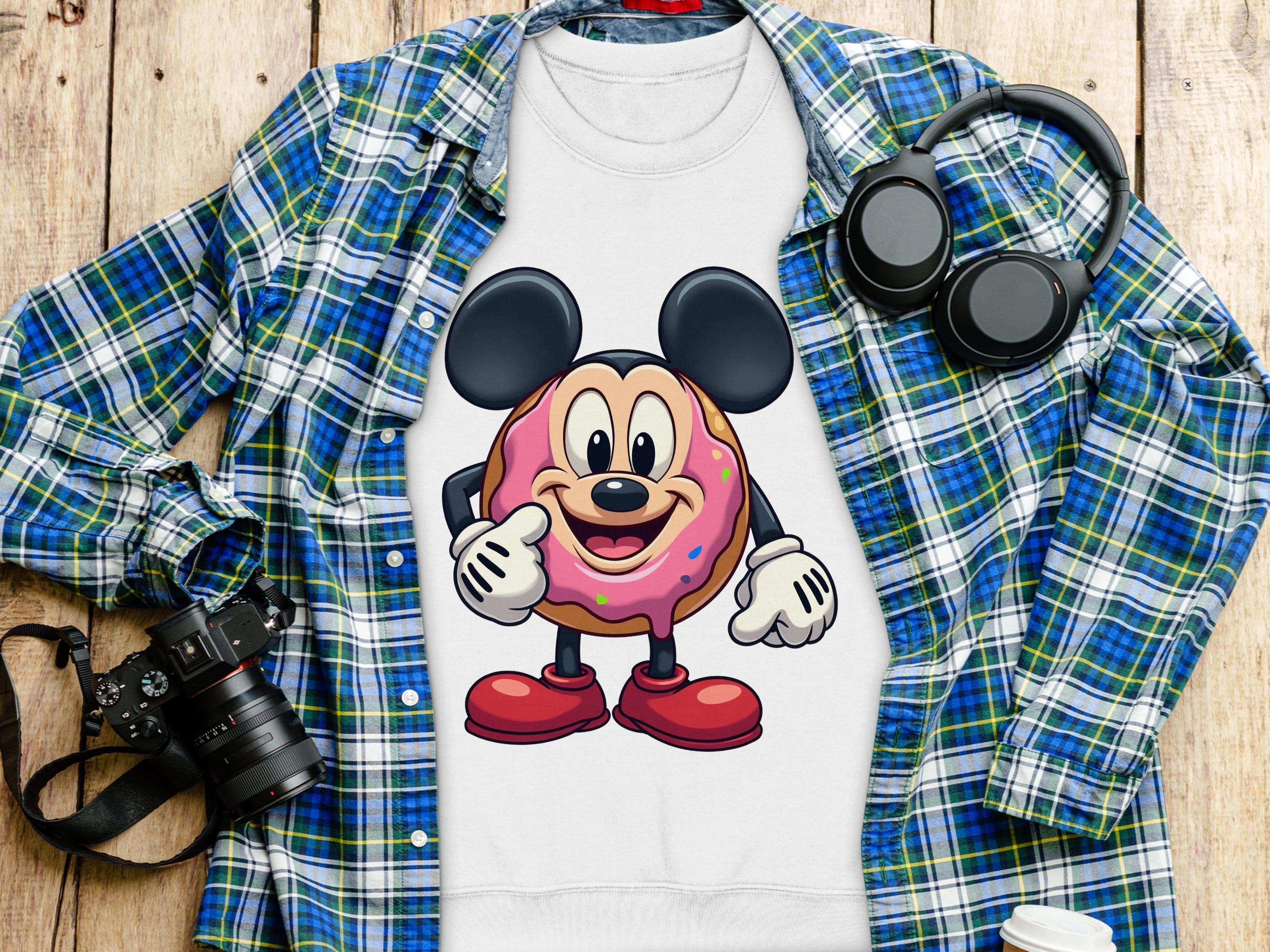 A quirky white T-shirt by Garment Graphics, featuring a cute cartoon mouse inside a donut, rests on a wooden surface. Nearby items for a playful wardrobe include a blue plaid shirt, headphones, a camera, and a white travel mug.