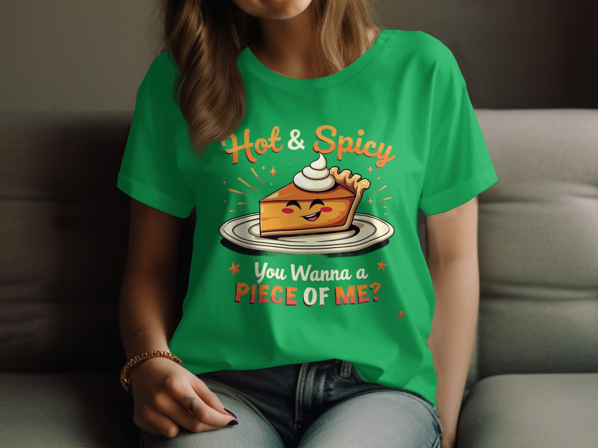 A woman sits on a sofa wearing a Garment Graphics green T-shirt, designed for casual outings in womens sizes. The shirt displays an illustrated pie slice with a face and whipped cream, humorously captioned Hot & Spicy, You Wanna a Piece of Me?.