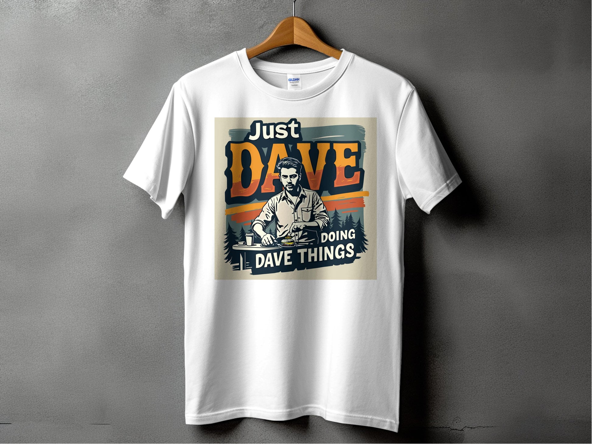 A classic white “Just Dave Doing Dave Things” graphic tee from Garment Graphics is displayed on a wooden hanger against a dark gray backdrop, featuring stylized text and a man’s image for an eye-catching look.