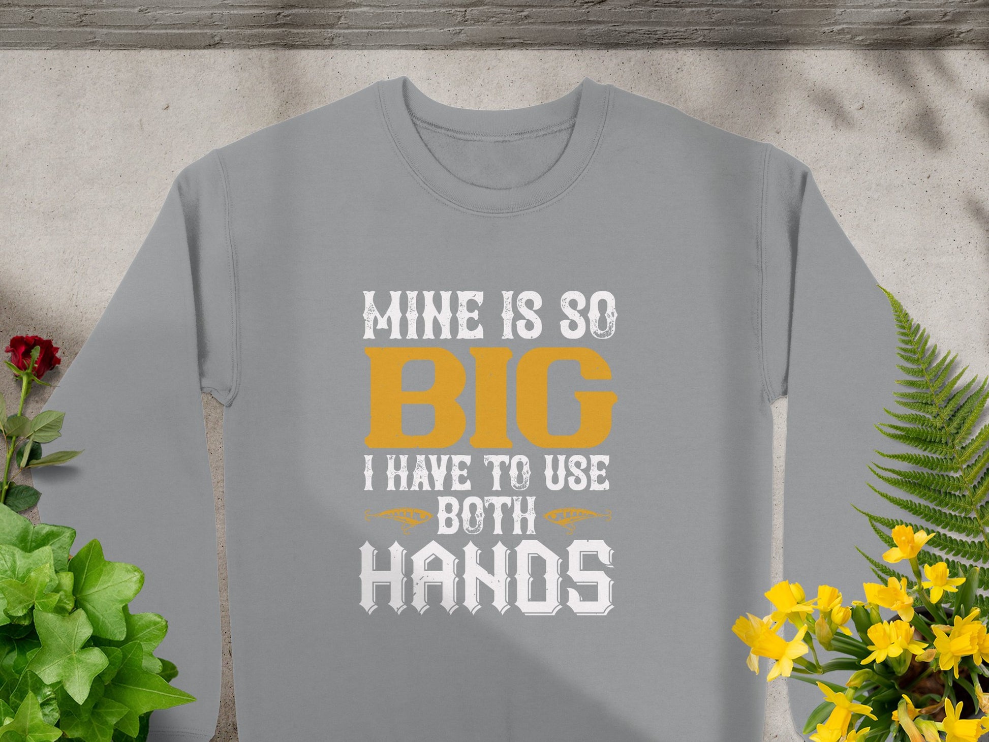 A Garment Graphics gray shirt boldly features Mine is so big I have to use both hands in yellow and white text, surrounded by green plants and vibrant yellow flowers, mirroring the daring style of unique sweatshirts.