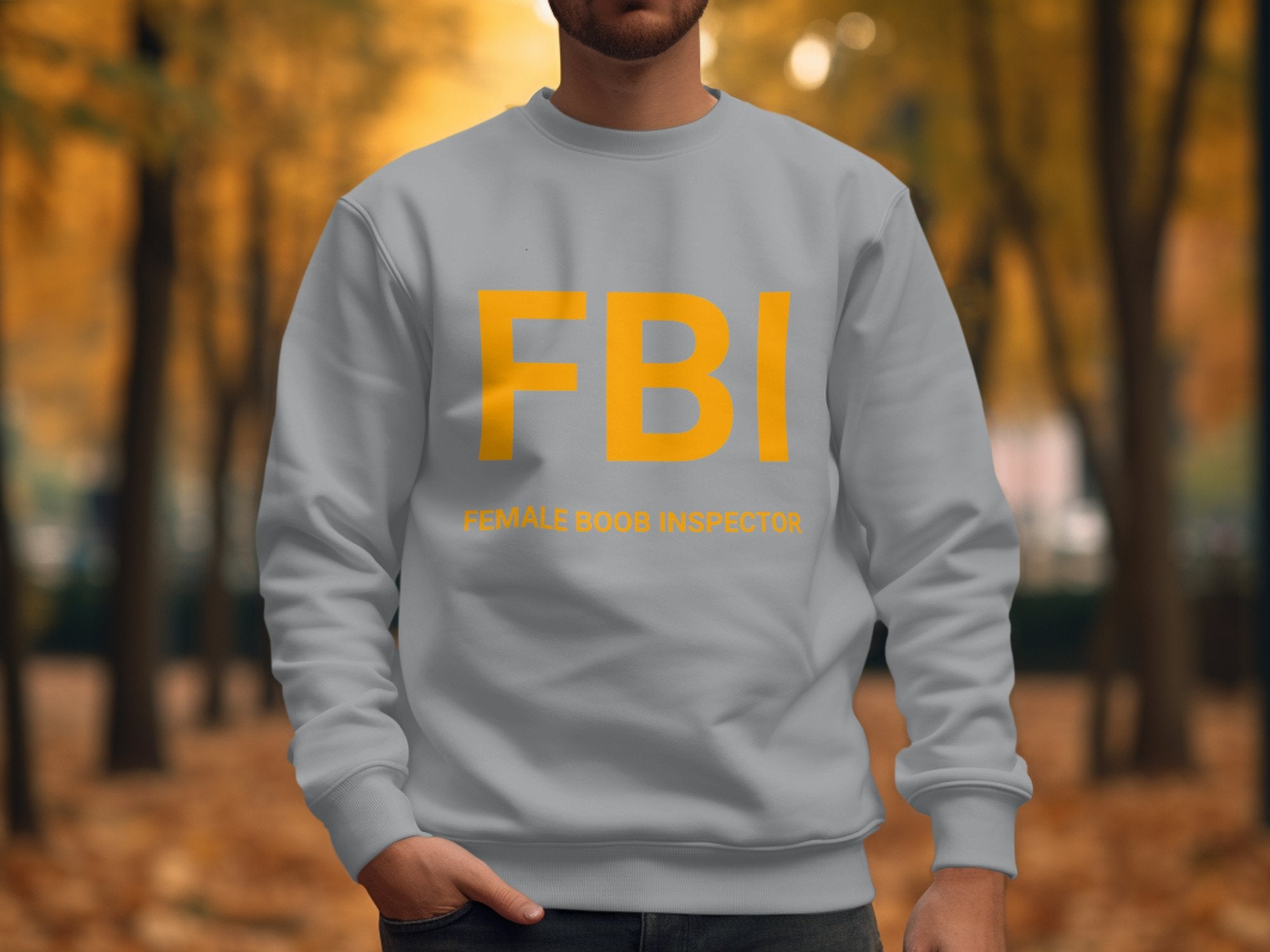 A person in Garment Graphics light gray FBI sweatshirt with yellow lettering enjoys an autumn walk. The smaller text humorously says Female Boob Inspector, showcasing the playful, unisex design against a warm, soft-focus backdrop of trees.