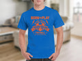 A person is wearing a bright blue football T-shirt from Garment Graphics featuring an orange design with a soccer ball and text BORN TO PLAY and YOUNG SQUAD FOOTBALL. The shirt subtly incorporates motivational phrases like One Goal One Dream in a blurred indoor setting.