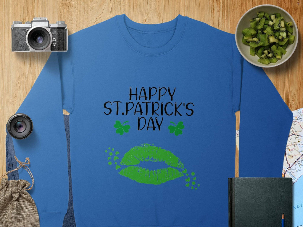 Garment Graphics blue sweatshirt, featuring Happy St. Patricks Day text and green lips graphic, captures the festive clover kiss spirit, displayed among a camera, lens, kiwi bowl, map, pouch, and closed notebook on a wooden surface.