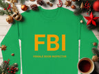 A unisex green sweatshirt from Garment Graphics features the yellow text FBI and Female Boob Inspector, displayed on a wooden surface. Pine branches, Christmas ornaments, a candy cane, and a cup of coffee enhance the festive vibe for quirky holiday fun.