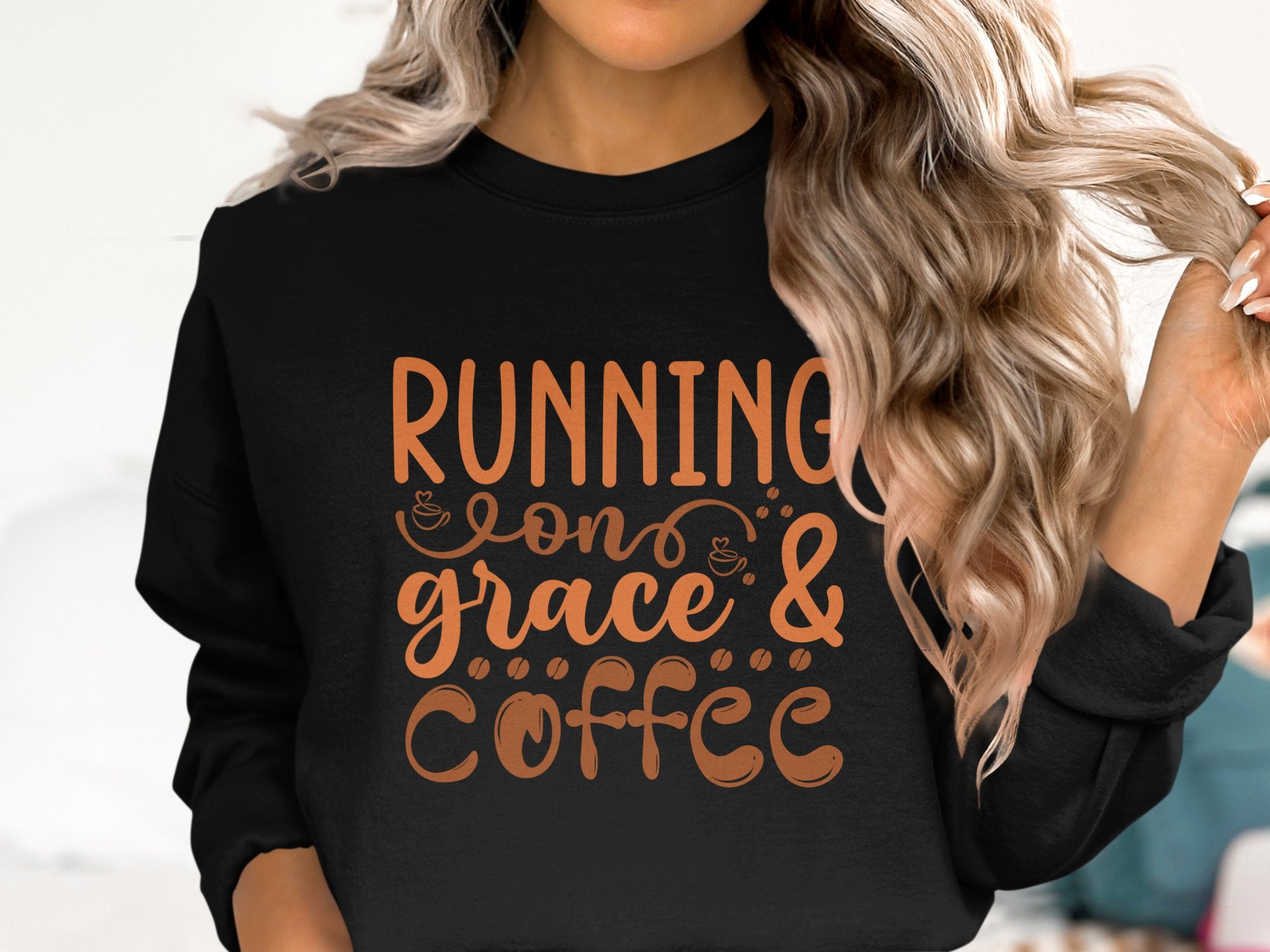 A person with long, wavy hair wears a Garment Graphics unisex graphic sweatshirt that says Running on Grace & Coffee in bold orange letters with playful coffee cup icons, embodying casual elegance.