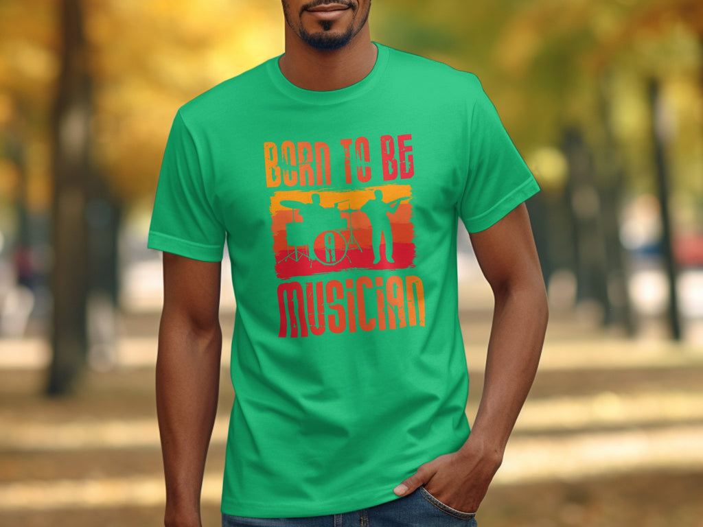 Someone is wearing a bright green Garment Graphics Born To Be Musician T-shirt featuring drummer and guitarist silhouettes with a drum set, set against an outdoor path framed by fall-colored trees—a delightful scene for music enthusiasts.