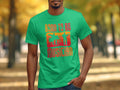 Someone is wearing a bright green Garment Graphics Born To Be Musician T-shirt featuring drummer and guitarist silhouettes with a drum set, set against an outdoor path framed by fall-colored trees—a delightful scene for music enthusiasts.
