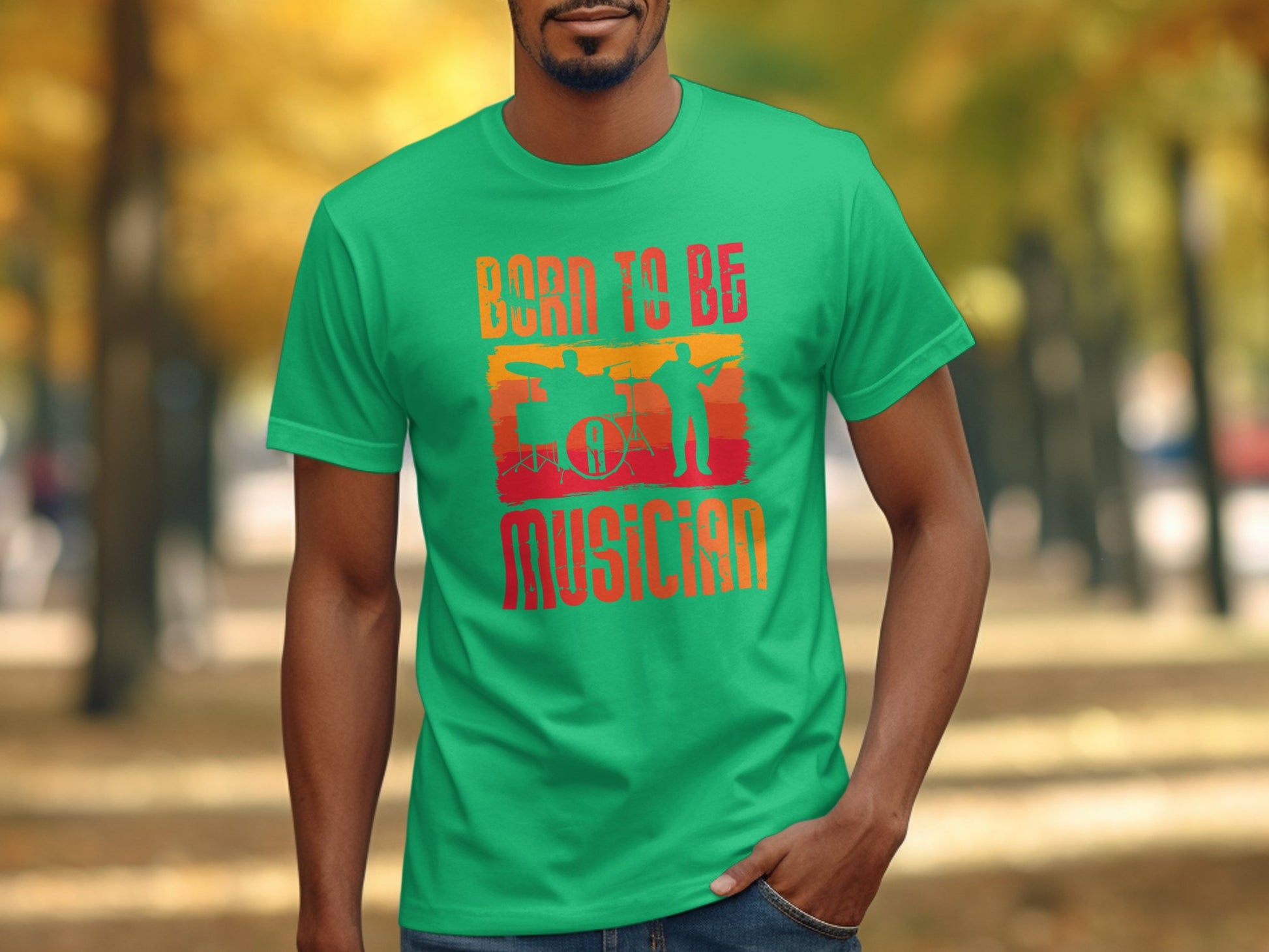 Someone is wearing a bright green Garment Graphics Born To Be Musician T-shirt featuring drummer and guitarist silhouettes with a drum set, set against an outdoor path framed by fall-colored trees—a delightful scene for music enthusiasts.