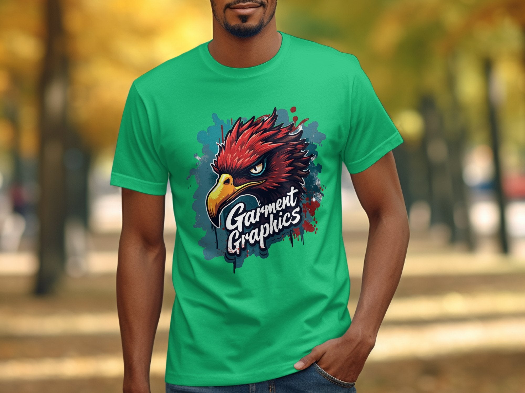 A person wears a green classic-fit T-shirt from Garment Graphics, featuring a red raptor head graphic and the brands name. A blurred pathway with autumn trees is in the background.