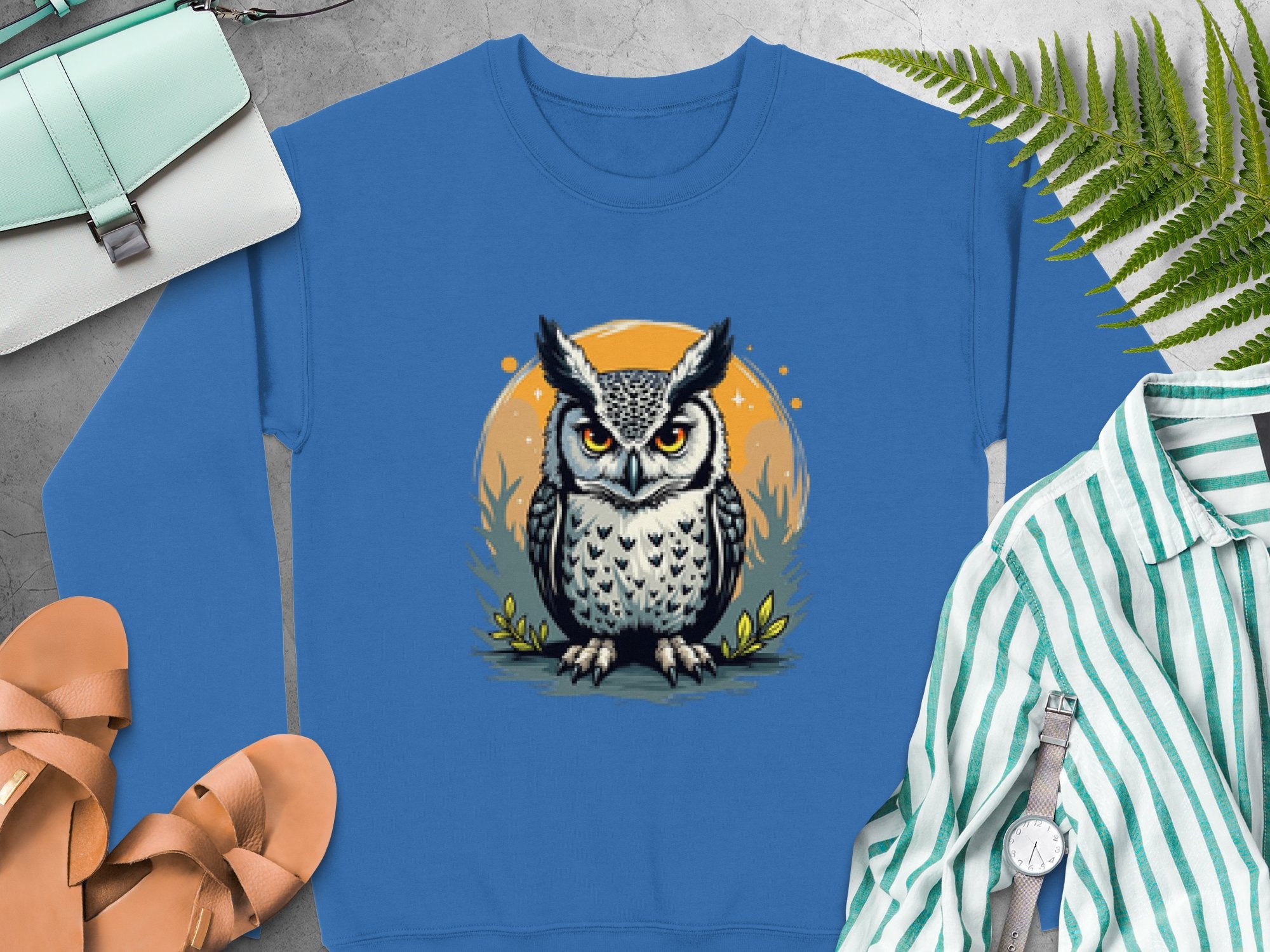 A unisex blue sweatshirt from Garment Graphics features an owl motif in gray within an orange circle. Its showcased with sandals, a mint purse, a striped shirt, a silver watch, and green ferns on a concrete background.
