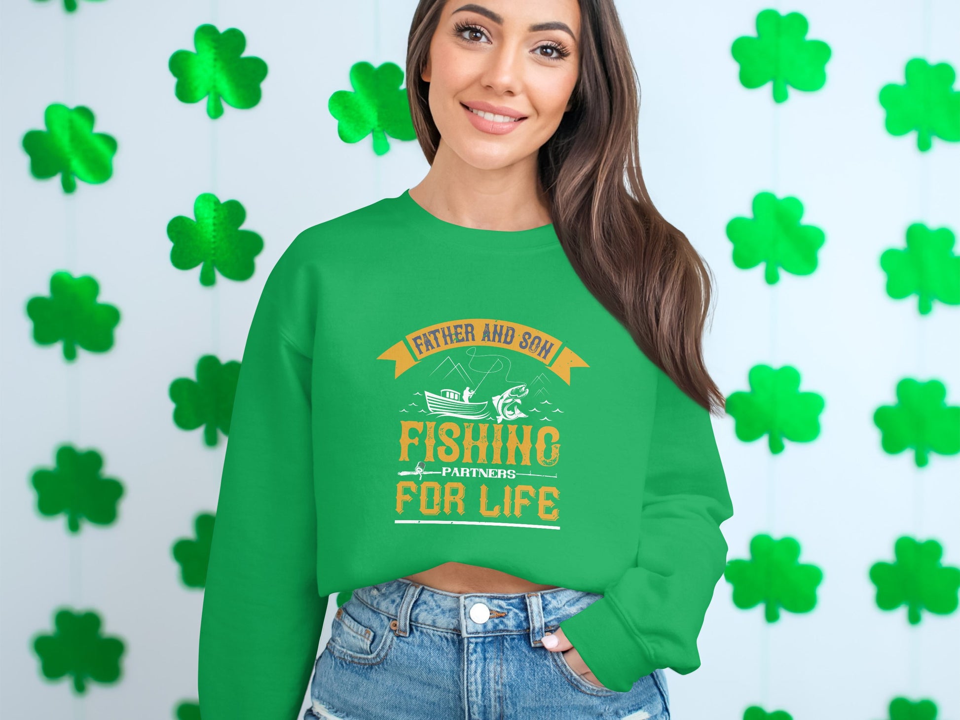 A woman stands against a white wall with green shamrocks, proudly wearing a Garment Graphics green sweatshirt that says Father and Son Fishing Partners for Life, featuring a charming fishing boat graphic.