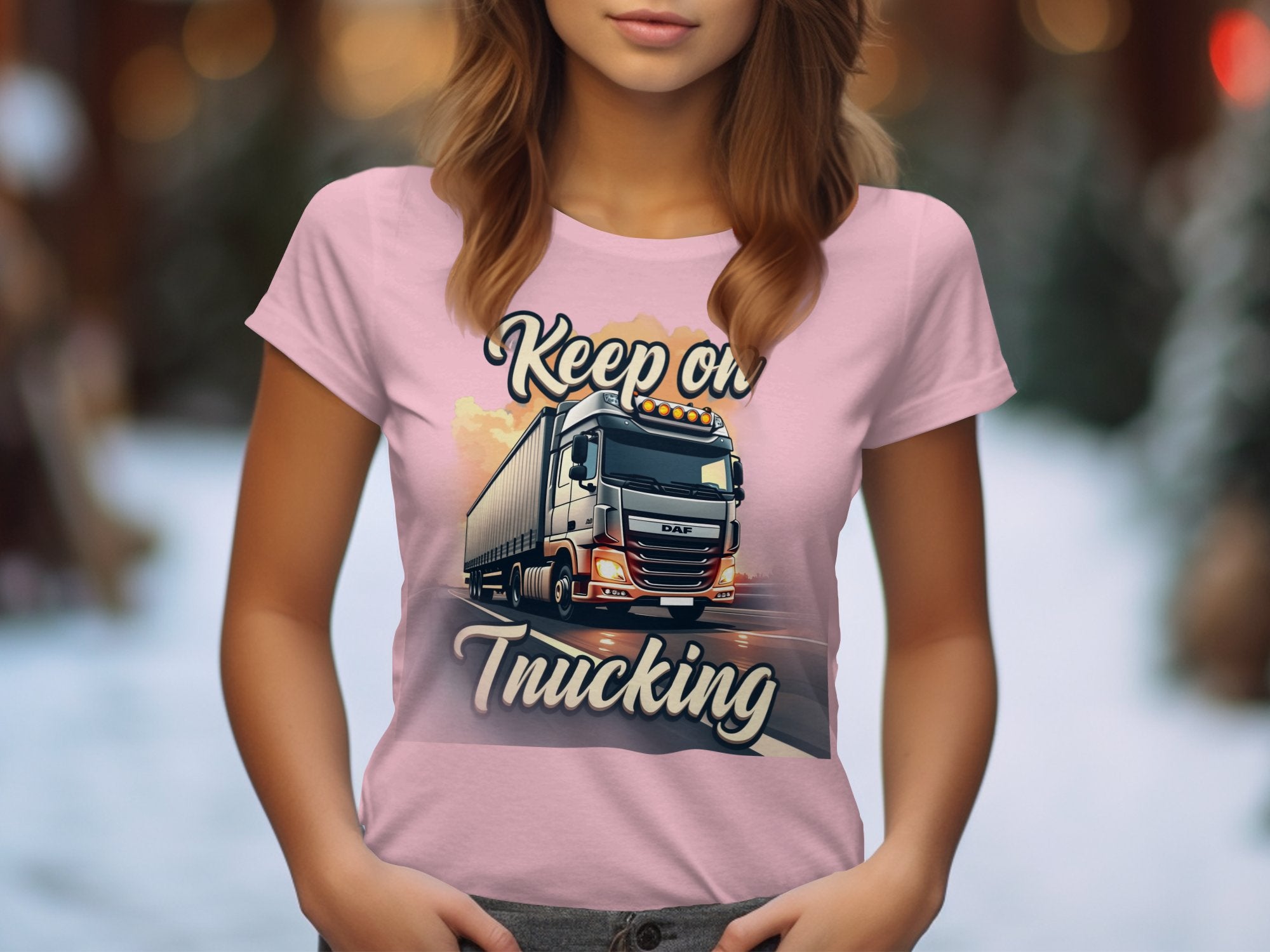 Wearing a pink graphic T-shirt by Garment Graphics, featuring a powerful truck and bold Keep on Trucking text, the person stands against a blurred background of warm lights and dark greens, suggesting an outdoor or festive atmosphere.