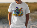 A man wears a Garment Graphics white t-shirt with vibrant graffiti-style art of two dancers and Risk Taker Street Dance 10th Final. He stands outdoors, embodying street dance culture against a backdrop of grass and sand with a blurred horizon.