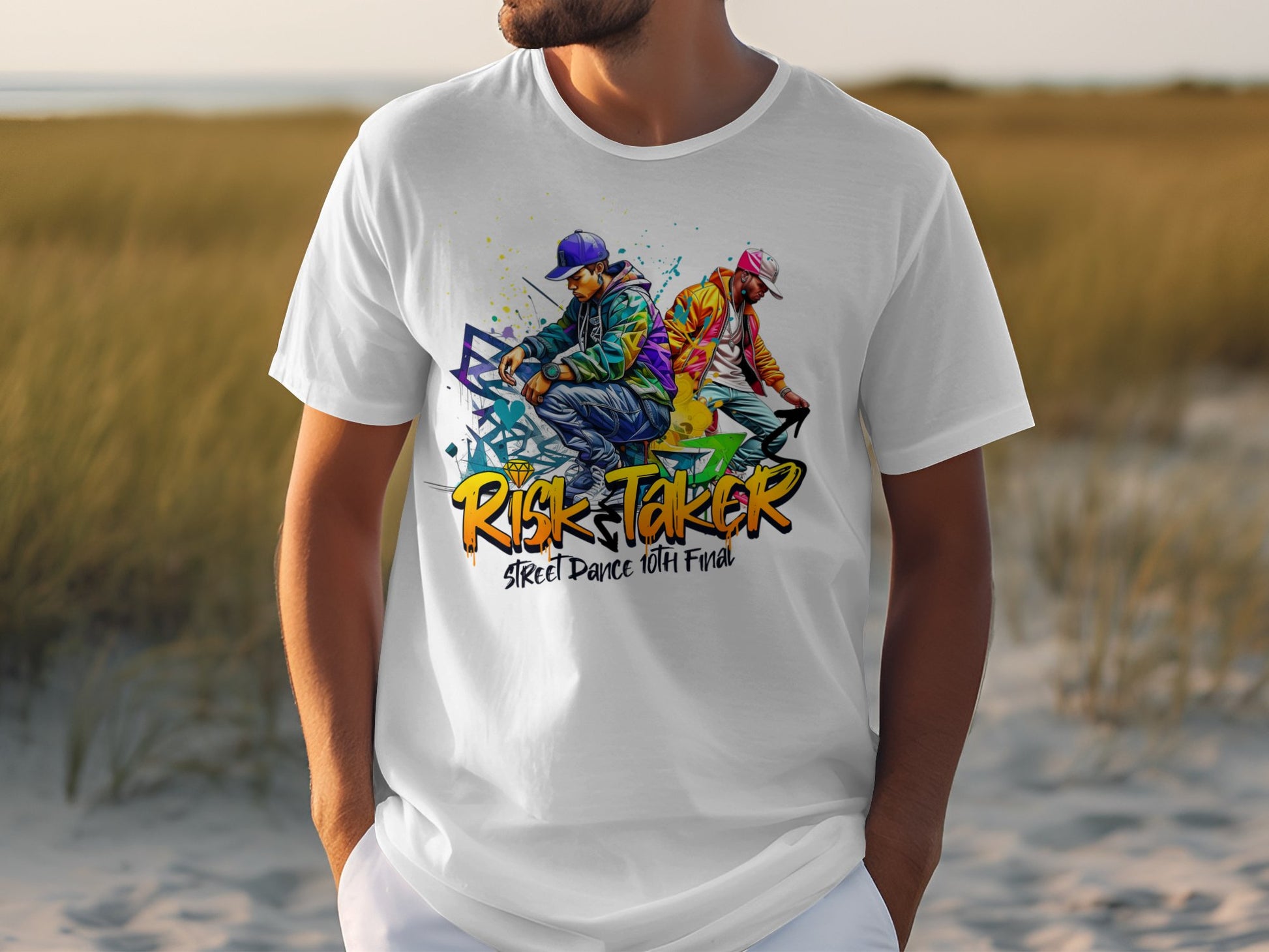 A man wears a Garment Graphics white t-shirt with vibrant graffiti-style art of two dancers and Risk Taker Street Dance 10th Final. He stands outdoors, embodying street dance culture against a backdrop of grass and sand with a blurred horizon.