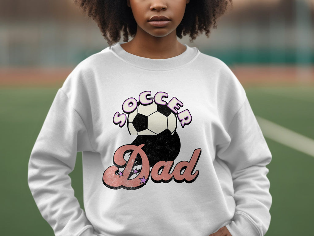 A person wearing a Garment Graphics white graphic print sweatshirt featuring Soccer Dad with a retro soccer ball design stands outdoors against a blurred green and brown backdrop.