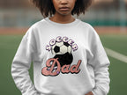 A person wearing a Garment Graphics white graphic print sweatshirt featuring Soccer Dad with a retro soccer ball design stands outdoors against a blurred green and brown backdrop.