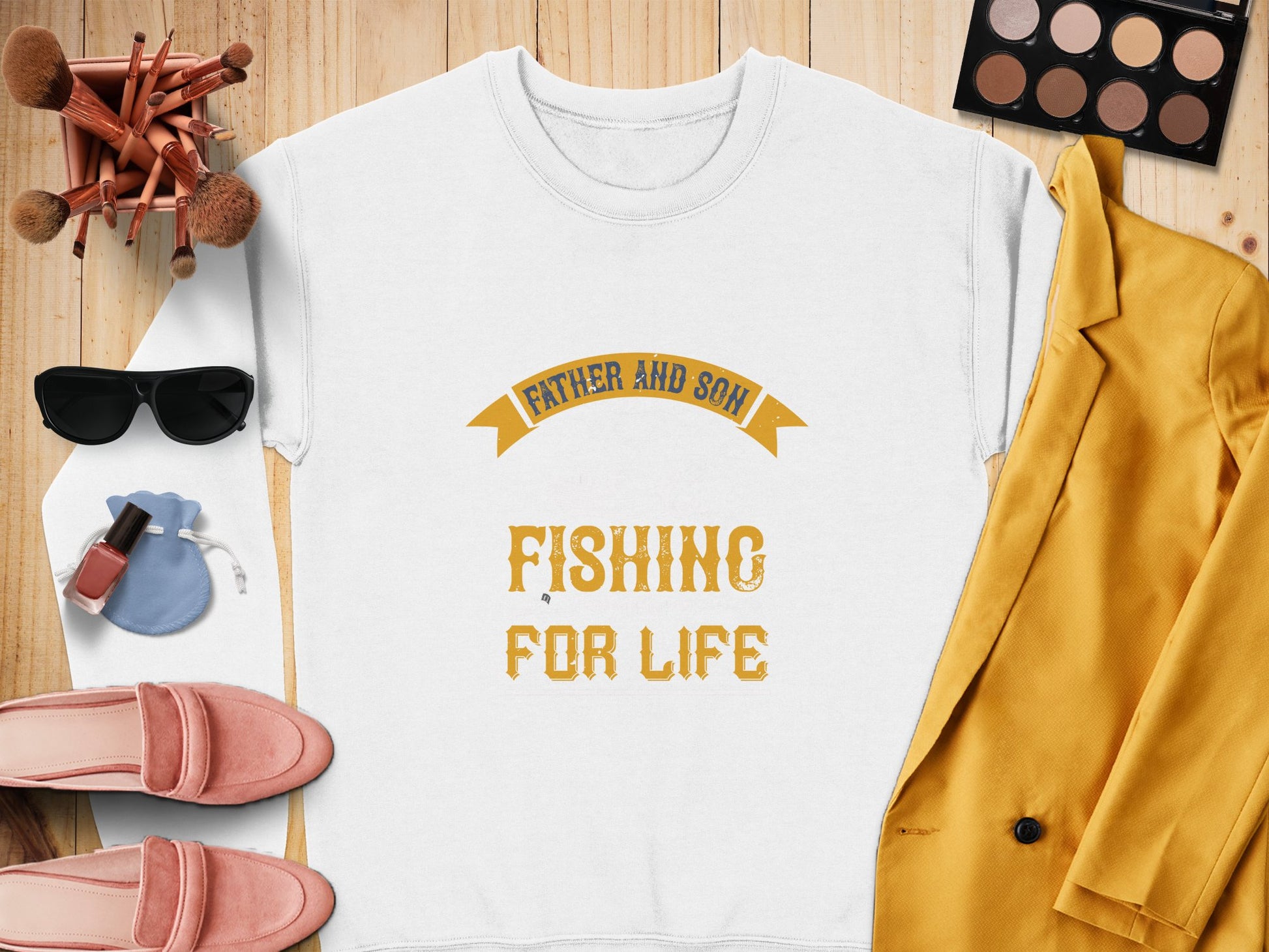 A Father and Son Fishing for Life white T-shirt by Garment Graphics, featuring yellow lettering, is surrounded by a mustard blazer, pink shoes, makeup brushes, an eyeshadow palette, black sunglasses, nail polish, and a blue drawstring pouch on a wooden surface—perfect fishing prep.