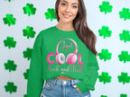 A woman sports a Garment Graphics green sweatshirt with Just be COOL, Rock and Roll! in pink and white, complemented by vibrant pink headphones. She poses against a light background decorated with charming green clovers, creating an ideal look for music enthusiasts.