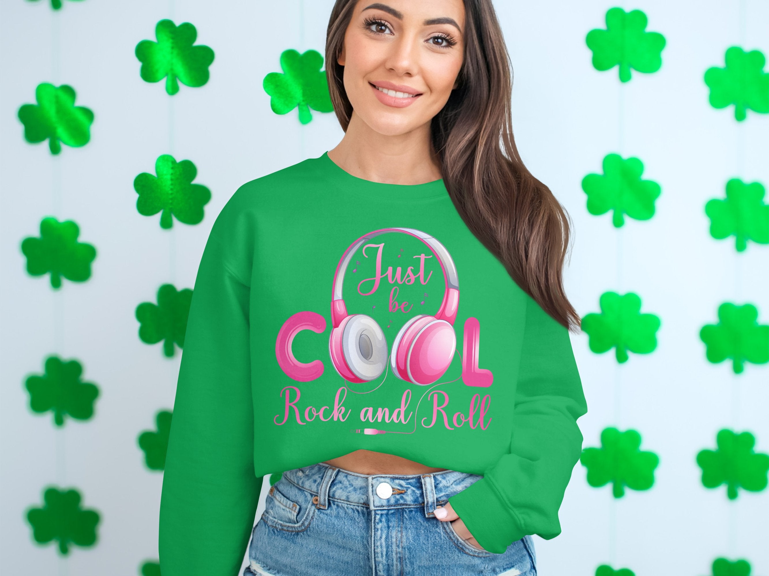 A woman sports a Garment Graphics green sweatshirt with Just be COOL, Rock and Roll! in pink and white, complemented by vibrant pink headphones. She poses against a light background decorated with charming green clovers, creating an ideal look for music enthusiasts.