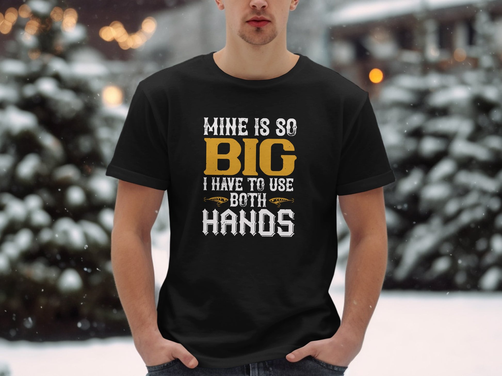 In a snowy outdoor scene, a person wears a black Garment Graphics T-shirt with the playful slogan Mine is so big I have to use both hands in white and orange. Perfect for fishing enthusiasts, the individual stands with their hands in their pockets.