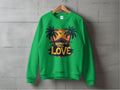 A green sweatshirt by Garment Graphics hangs on a wooden hanger against a concrete wall, featuring a vibrant sunset between palm trees and bold One Love lettering. Unisex sizing ensures this One Love Sweatshirt embodies effortless style and comfort.