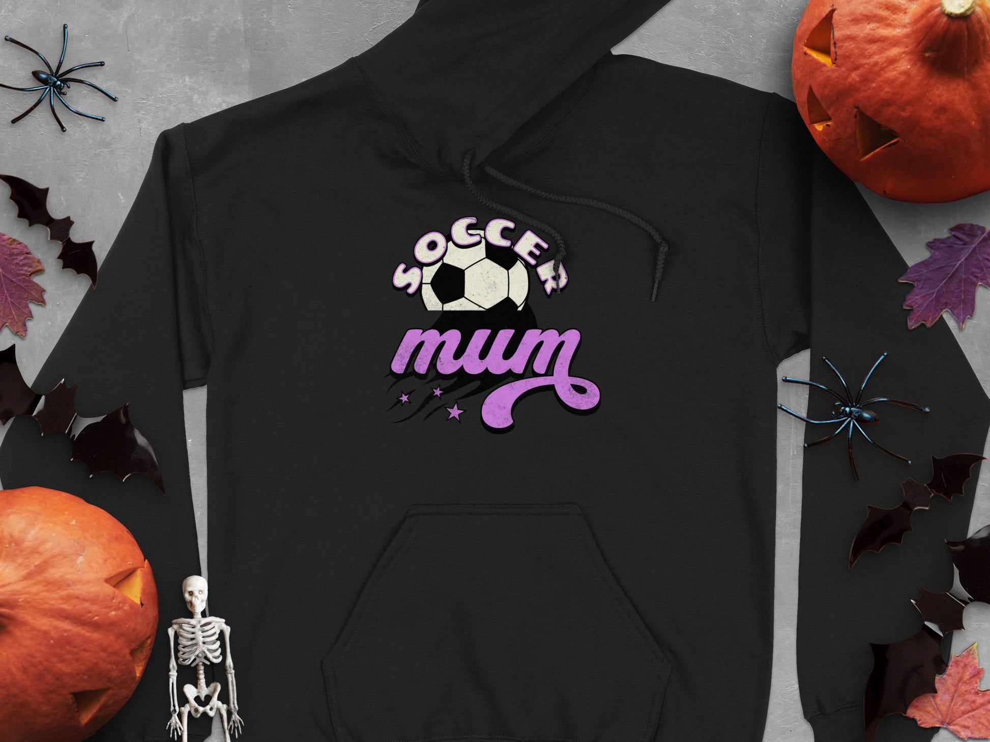 The Garment Graphics Soccer Mum Hoodie provides warmth and comfort with its Soccer Mum text and soccer ball graphic. Ideal for Halloween, it complements classic decorations like pumpkins, bats, skeletons, and leaves on a gray background. Classic fit ensures ultimate comfort!.