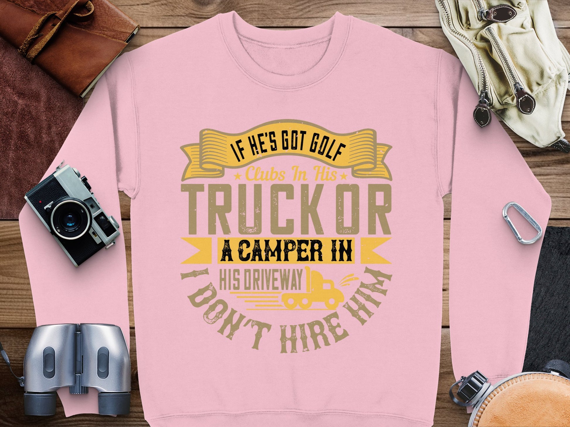A Garment Graphics pink sweatshirt with the cheeky phrase: If hes got golf clubs in his truck or a camper in his driveway, I dont hire him, rests on wood. Nearby are a camera, binoculars, pouch, and gloves—ideal for those who love casual wear with humor.