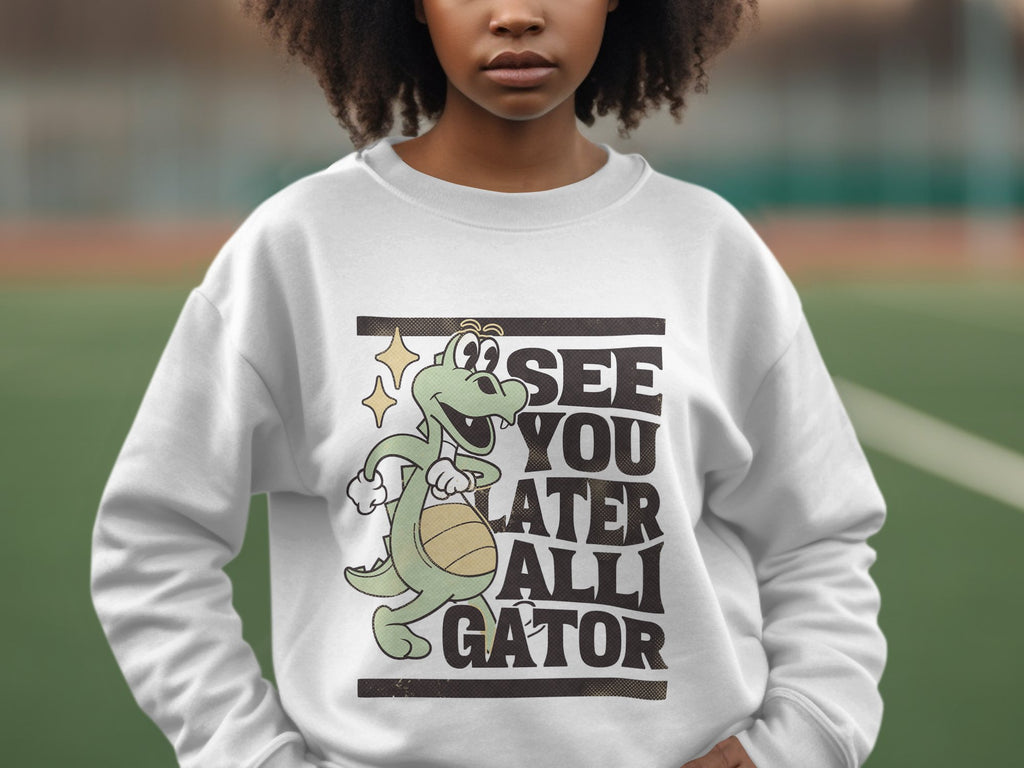 A person stands outdoors wearing a Garment Graphics white sweatshirt featuring a playful alligator holding a basketball with the text, See You Later Alligator. The blurred background highlights the sweatshirts fun design.