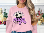 In a kitchen, someone wears a Garment Graphics light pink sweatshirt featuring SOCCER Mum and a vintage soccer ball graphic. Their long, wavy blonde hair flows gracefully as they pose with blurred holiday decorations in the background.