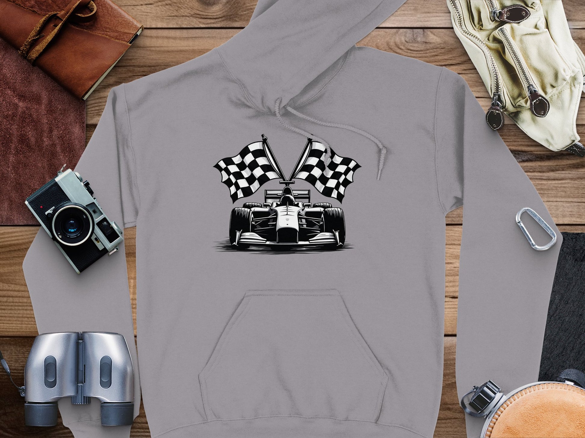 A Garment Graphics gray hoodie features a black and white race car with checkered flags, surrounded by binoculars, an old camera, a leather-bound book, a carabiner, and gloves on a wooden surface.