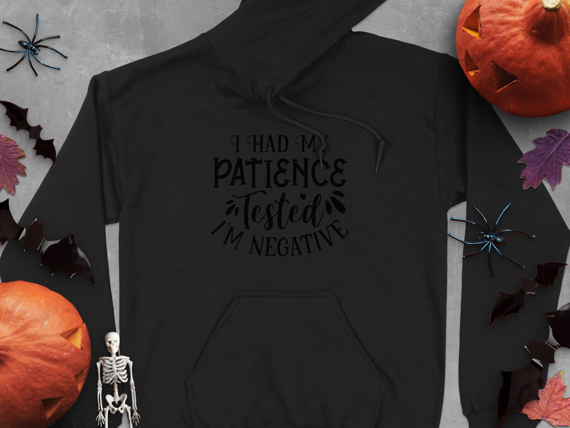 The Garment Graphics black hoodie says, I had my patience tested. Im negative, and is surrounded by Halloween decor—pumpkins, bats, a skeleton, and spiders on gray—a perfect choice for fans of unique, humorous statement hoodies.